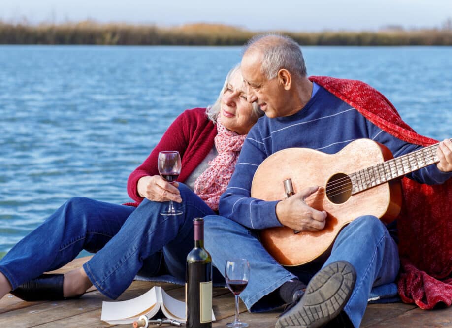 Happy senior couple enjoying time together playing guitar and drinking wine by the lake wrap around in a red blanket. Cottage Living.