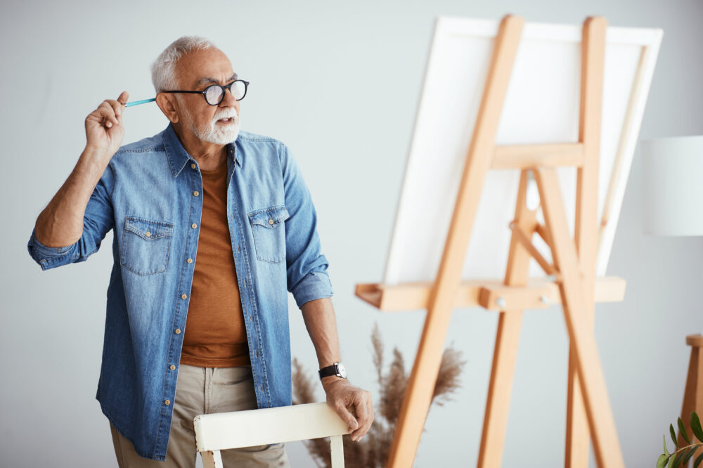 Independent living. Pensive senior man looking at his drawing on canvas while painting.