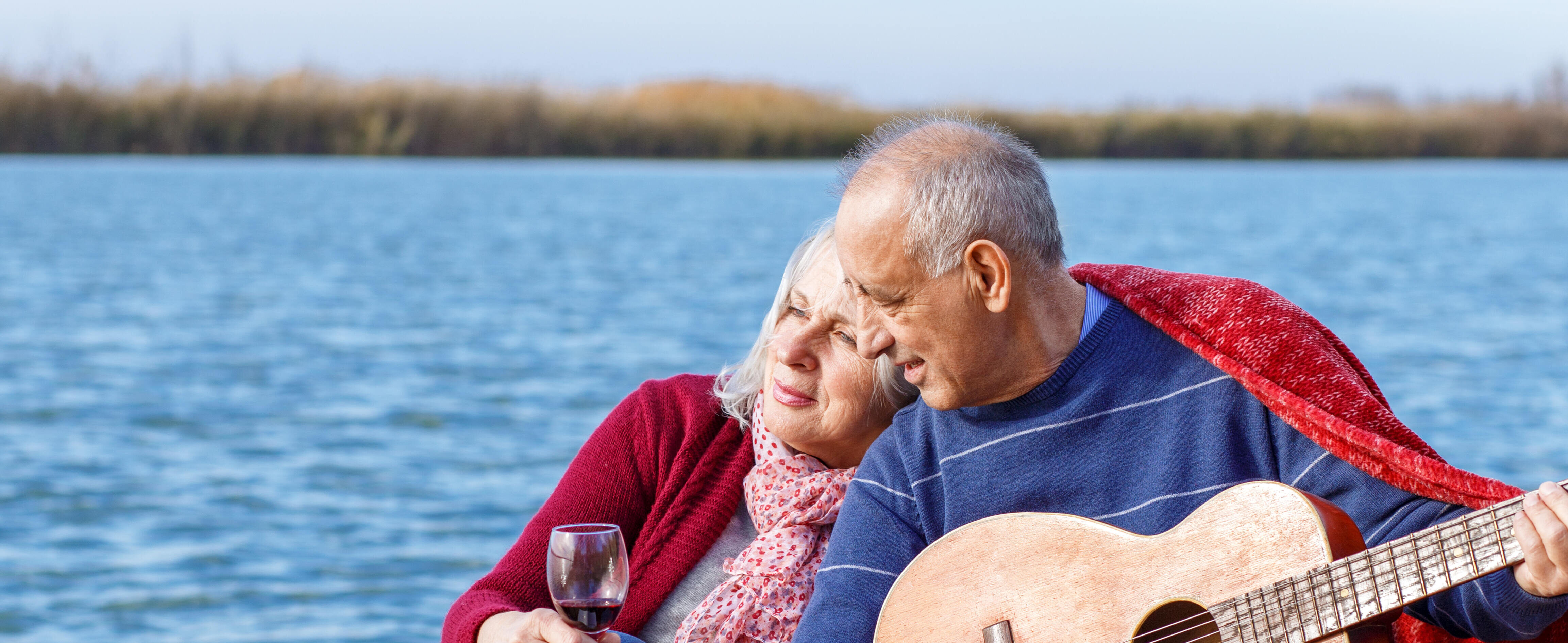 Happy senior couple enjoying time together playing the guitar and drinking wine by the lake.